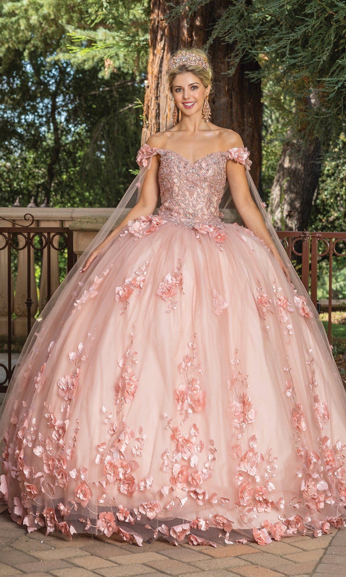 A woman stands outdoors wearing the Quinceanera Dress 1695 By Dancing Queen, a pink off-shoulder A-line gown with floral appliqués and a tiara, with trees and a fence in the background.
